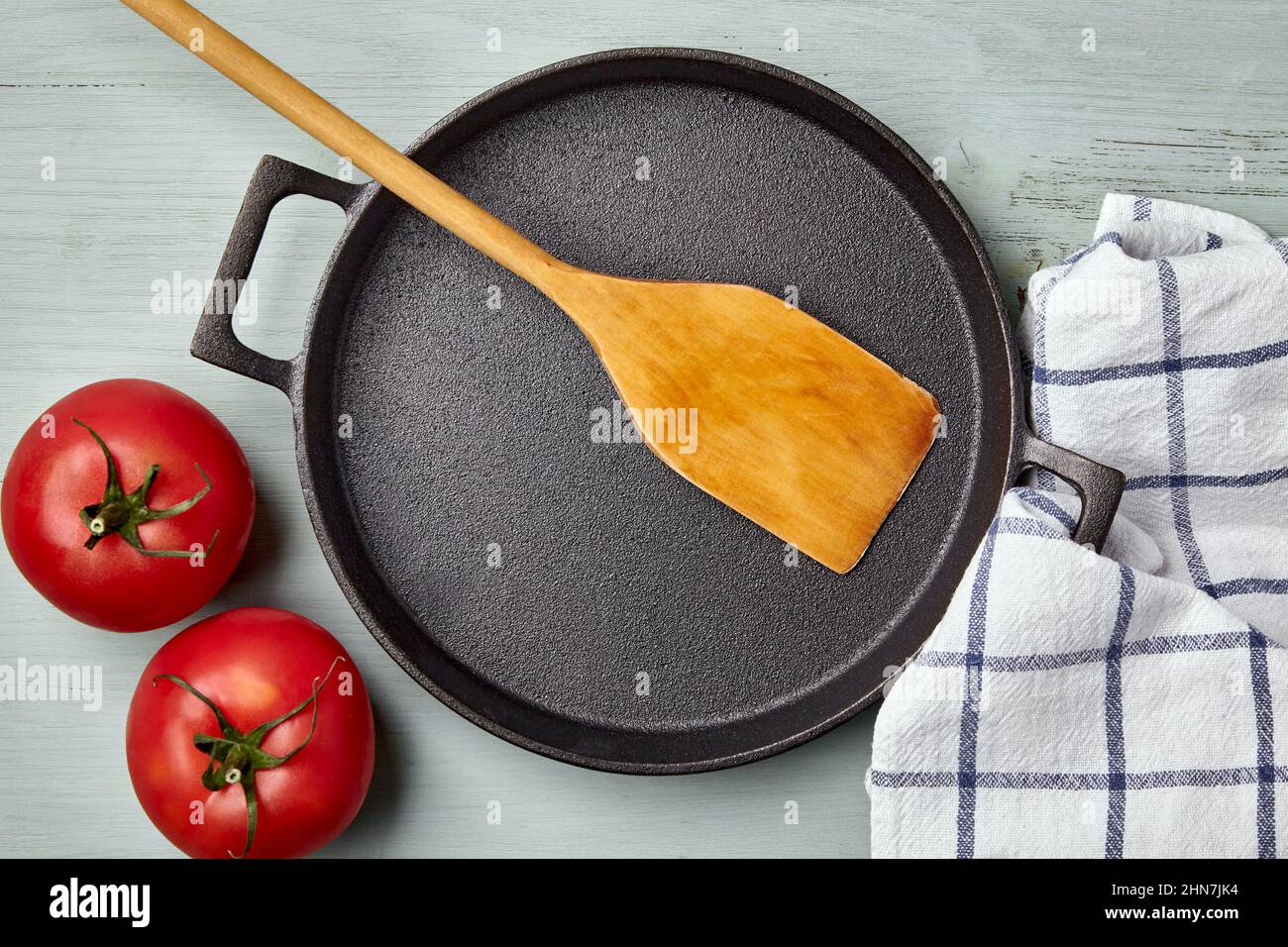Empty round cast iron pan, wooden spatula, tea towel and tomatoes Stock Photo