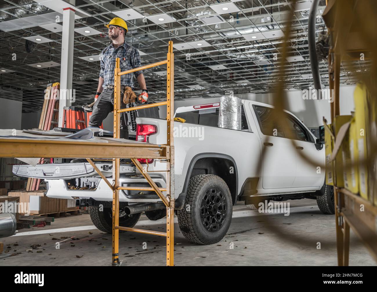 Caucasian Commercial Building Construction Worker in His 30s Wearing Hard Hat and Keeping Tools in His Hands Staying on His Pickup Truck Cargo Bed Stock Photo