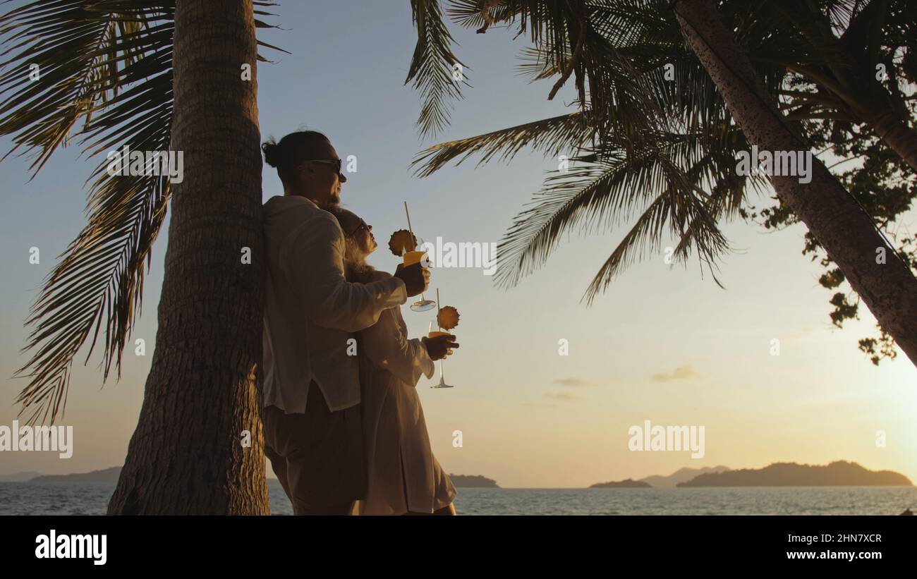 SILHOUETTE DARK GOLD SUNSET VIEW: Loving couple in white shirt and sunglasses, near palm tree, kissing, hugging, relax and drinking cocktail. Concept tropical summer vacations, holidays wedding love Stock Photo