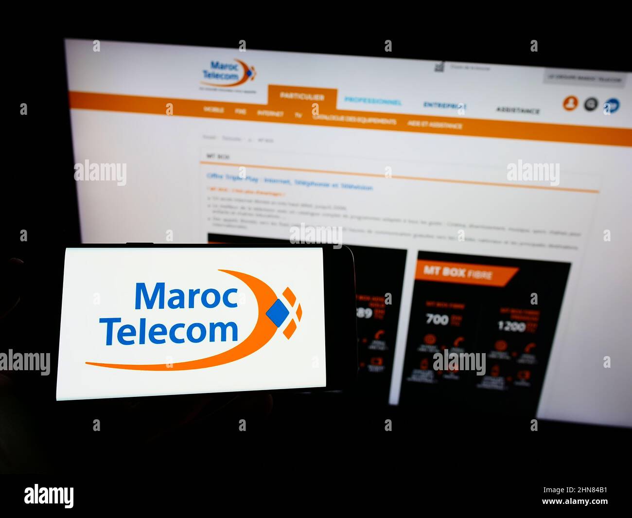 Person holding cellphone with logo of telecommunications company Maroc Telecom (IAM) on screen in front of webpage. Focus on phone display. Stock Photo