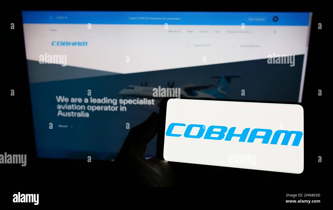 Person holding smartphone with logo of British aerospace company Cobham Limited on screen in front of website. Focus on phone display. Stock Photo