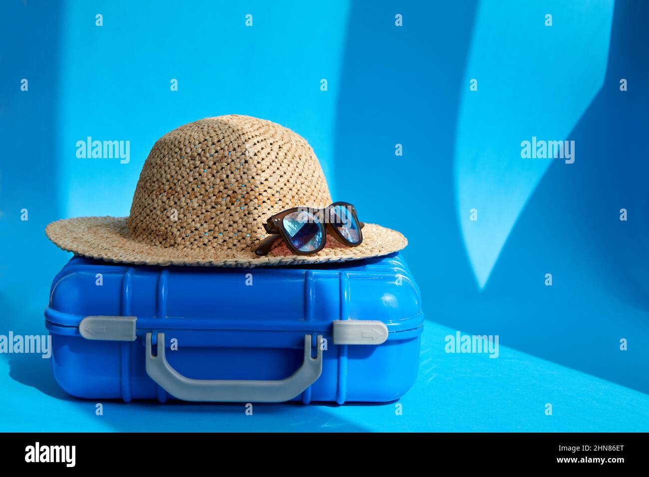 Plastic blue suitcase with straw hat and sunglasses placed on floor in bright studio for summer vacation Stock Photo