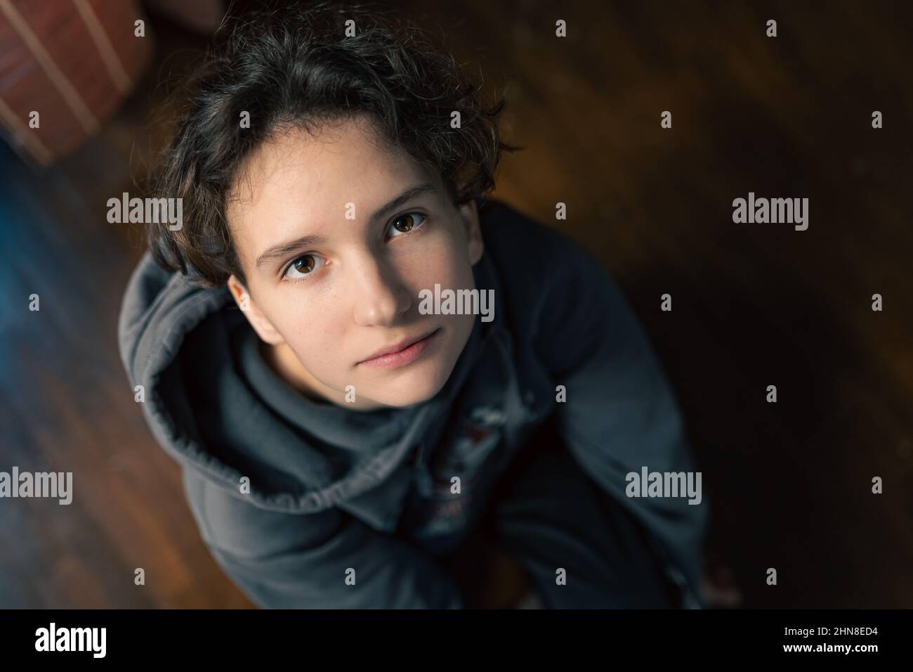 Portrait of teen girl with brown eyes looking at camera, brown curly hair sitting on floor. Looking up Stock Photo