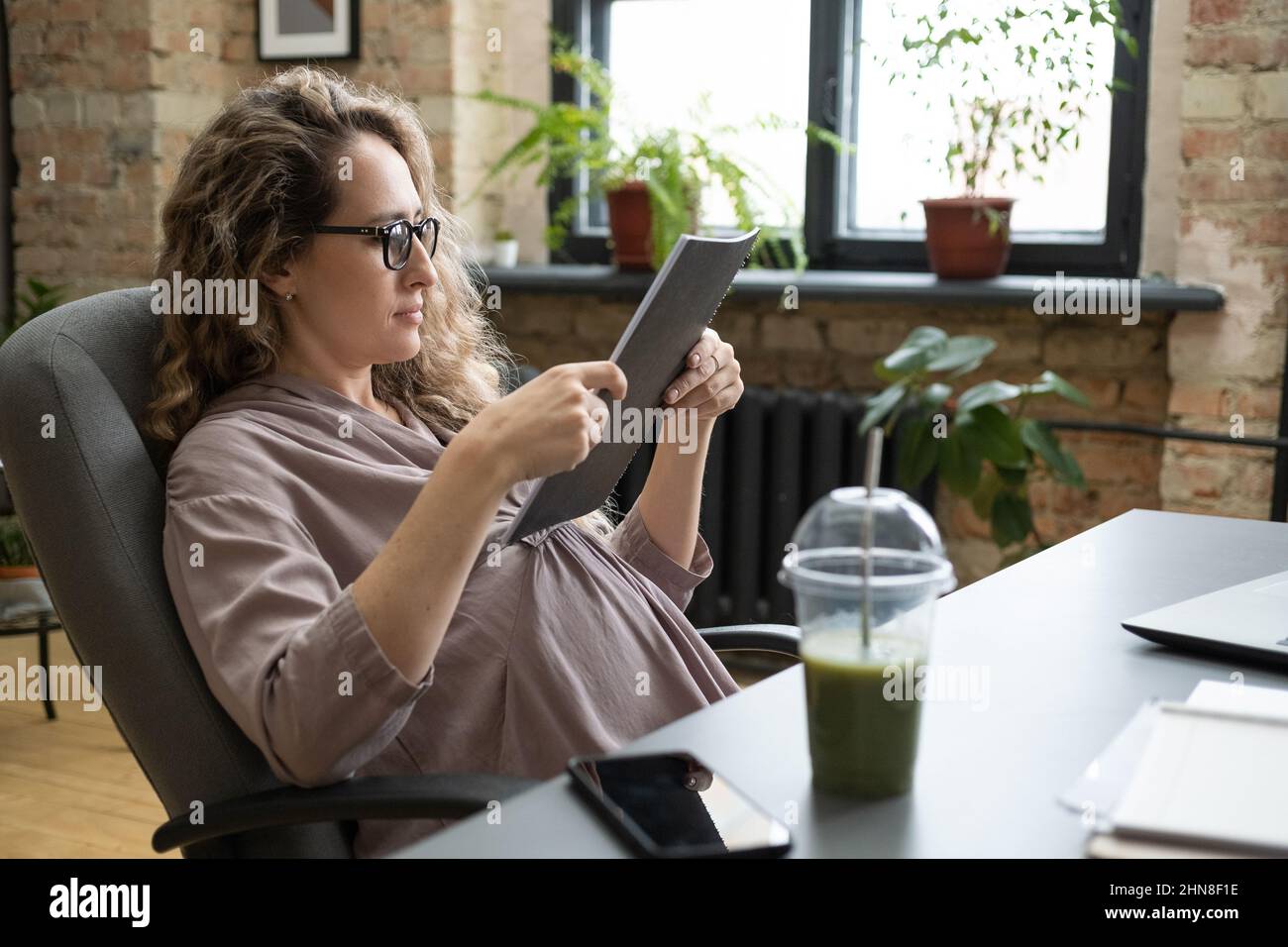 Pregnant businesswoman sitting on chair and reading documents in folder while sitting at the table with fresh cocktail Stock Photo