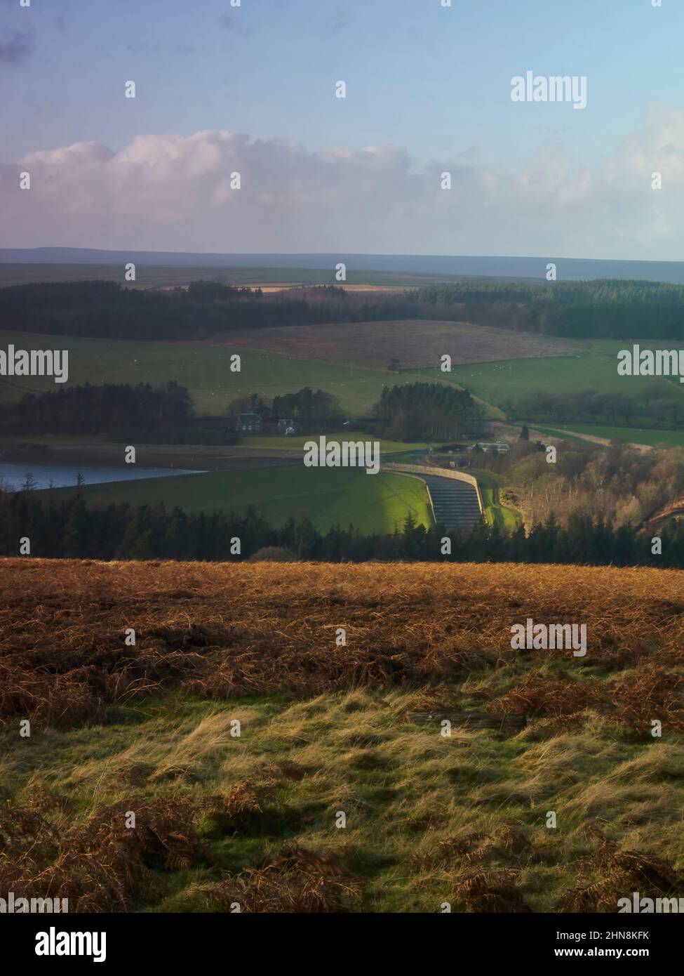 Clouds cast shadows over moorland, wooded hills and a reservoir spillway, all lit up with rich, golden winter sunlight. Stock Photo