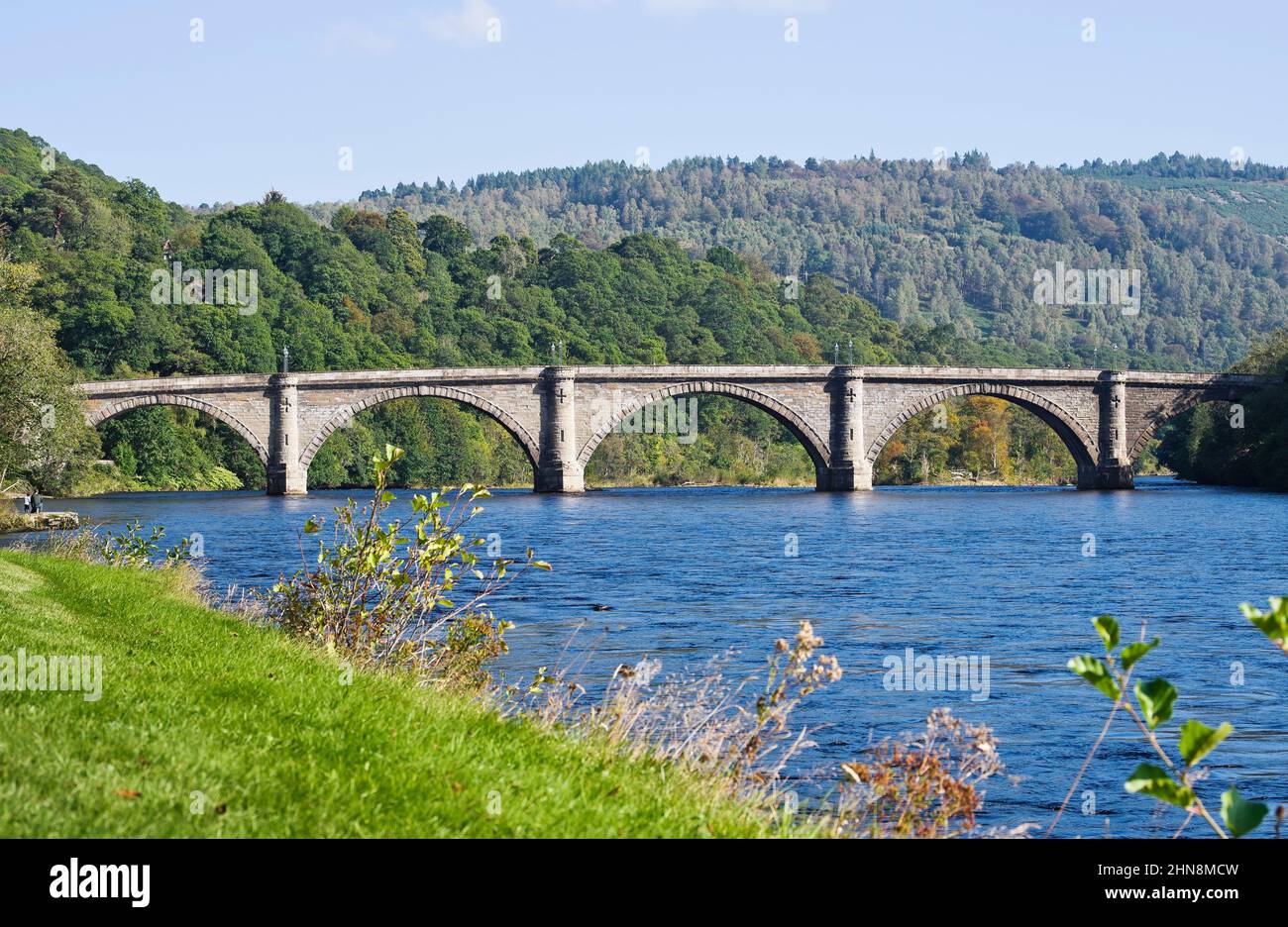 The old arched bridge over the River Tay at Dunkeld, Perthshire, Scotland, with thickly wooded hillsides forming a colourful green backdrop. Stock Photo