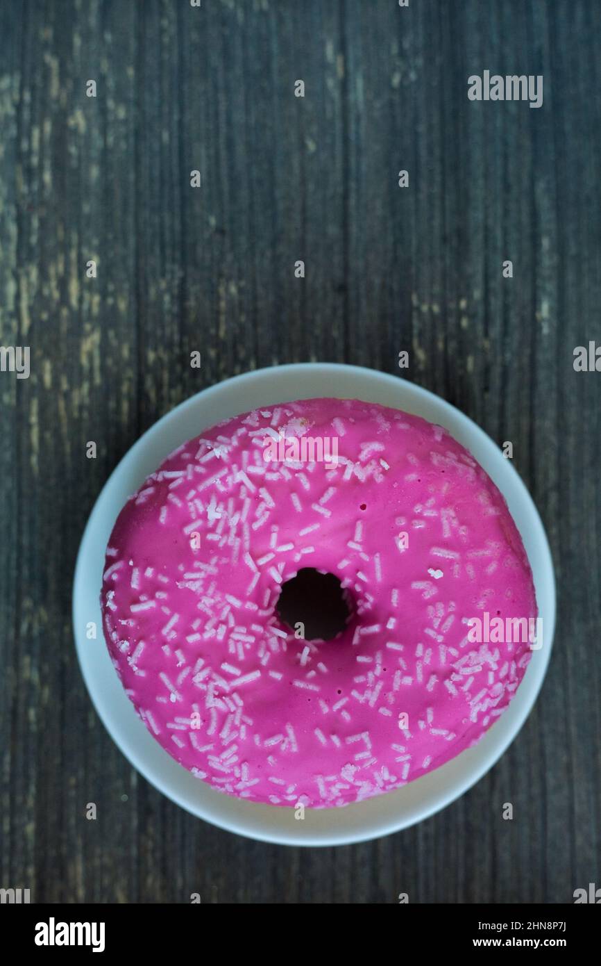 Vertical picture of bright pink donut on white round plate. Aged wooden background. Isolated object close up. Traditional American sweets for Stock Photo