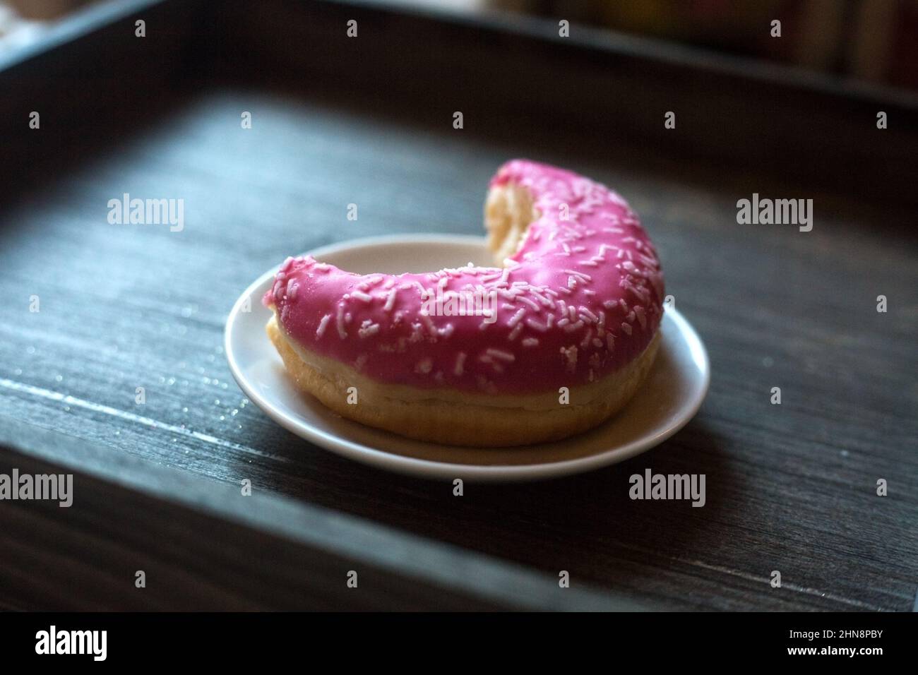 Side view to half eaten bright pink donut on white round plate. Aged wooden background. Isolated object. Traditional American sweets for breakfast Stock Photo