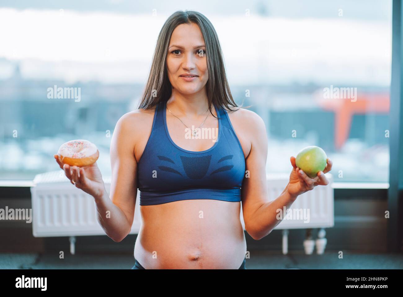 Fitness attractive pregnant woman in sportswear. Doing yoga exercise, choise green apple, donut. Diet starving. Close up Stock Photo