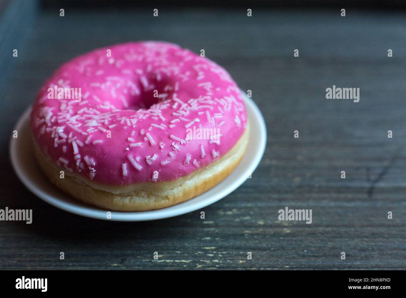 side view to bright pink donut on white round plate and aged wooden grey background. Isolated object close up. Traditional American sweets for Stock Photo