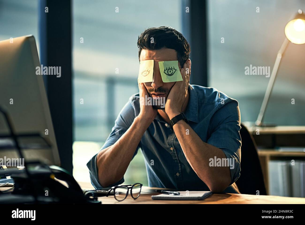 No time to sleep. Shot of a tired young businessman working late in an office with adhesive notes covering his eyes. Stock Photo