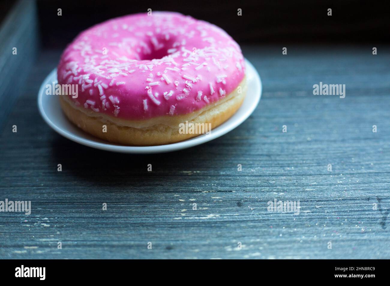 side view to bright pink donut on white round plate and aged wooden grey background. Isolated object close up. Traditional American sweets for Stock Photo