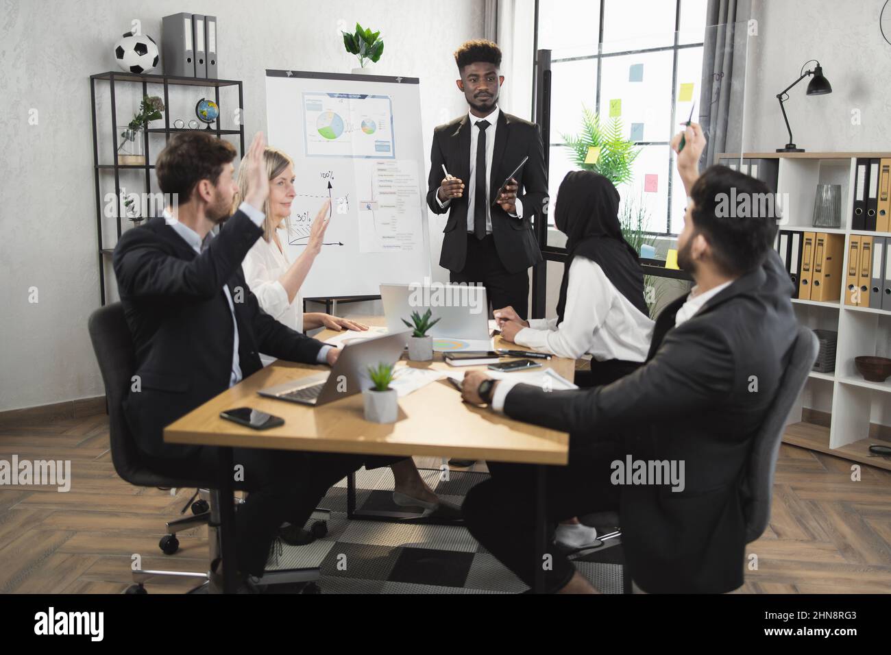 Multicultural business people in formal wear voting for some project during conference at office. Men and women sitting at desk with modern gadgets and raising hands. Stock Photo