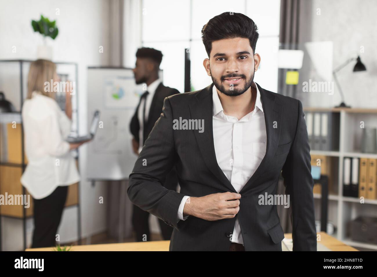 Successful indian man in suit smiling and looking at camera while his multi  ethnic colleagues talking on background. Office room for negotiation Stock  Photo - Alamy