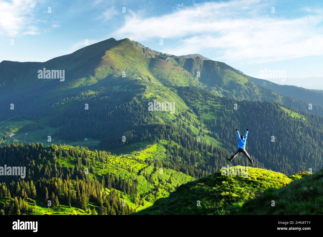 A tourist jump on the edge of a mountain covered with a lush grass. Purple sunset sky and hight mountains peaks on background. Landscape photography Stock Photo