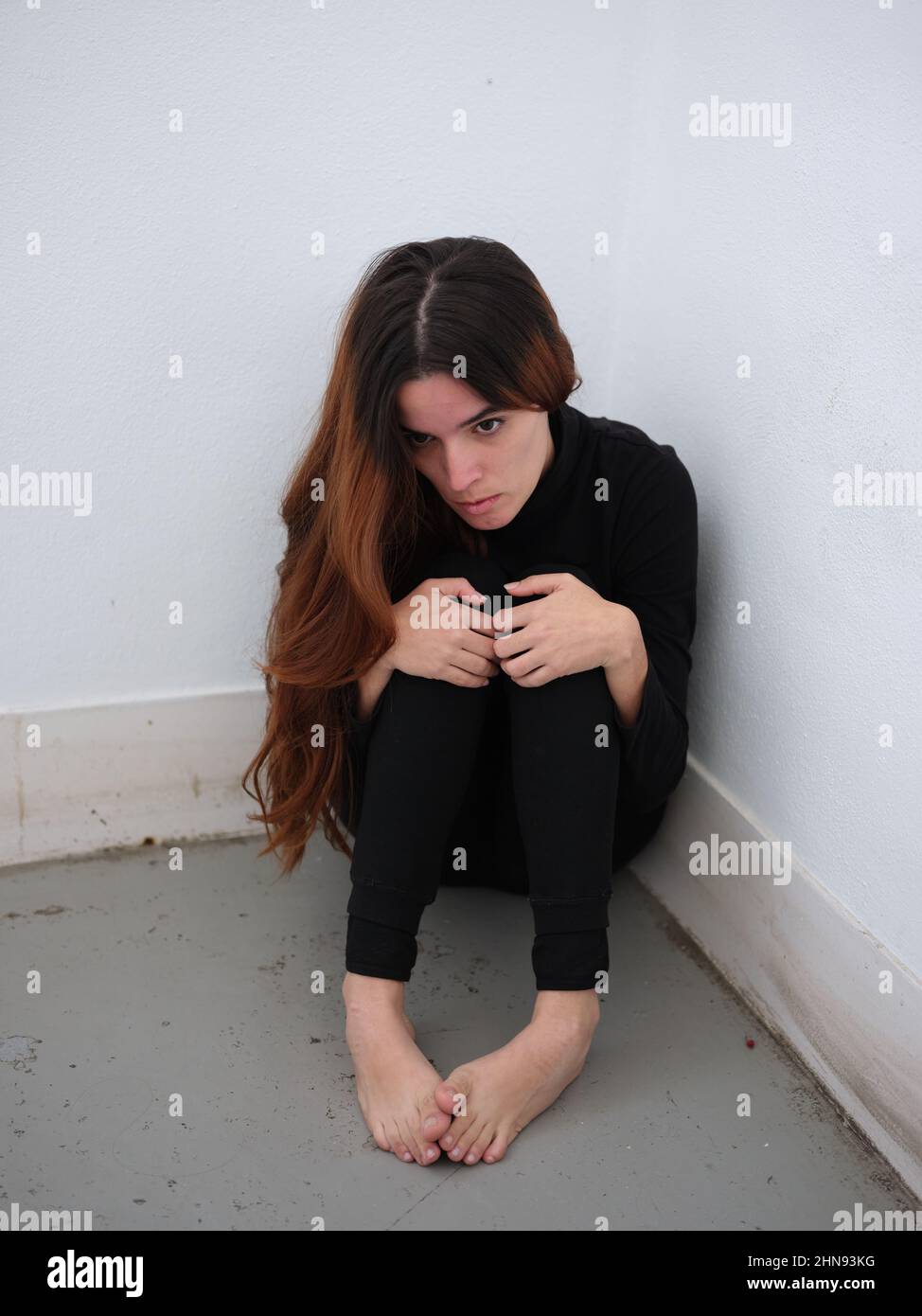 A young woman in the corner of a room dressed in black barefoot looking down in helplessly. Stock Photo