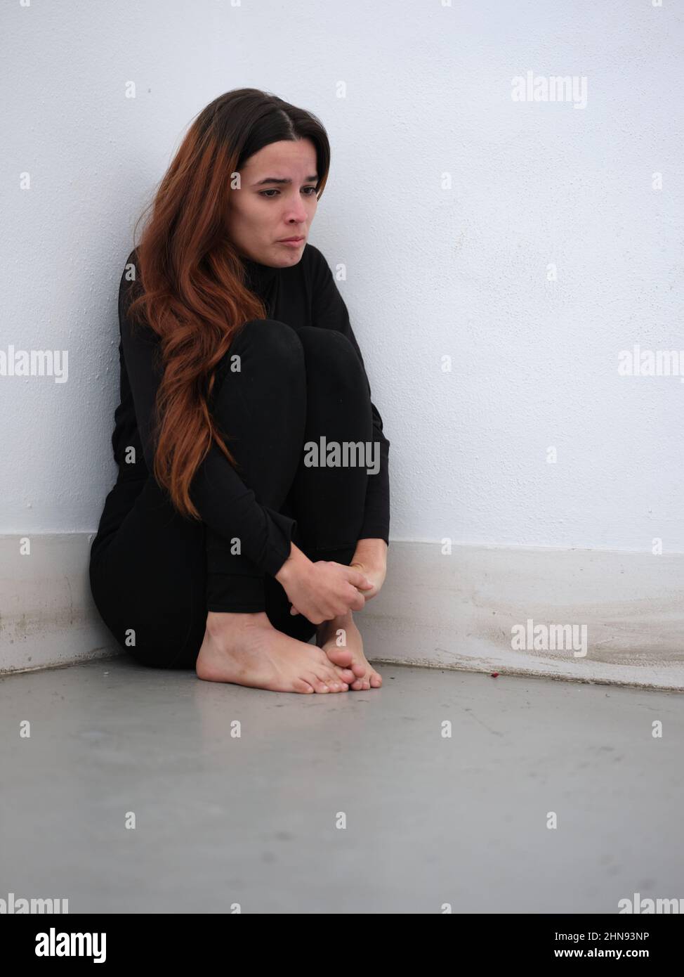 Low angle view of a sad young woman in the corner of a room dressed in black barefoot and on the verge of tears. Stock Photo