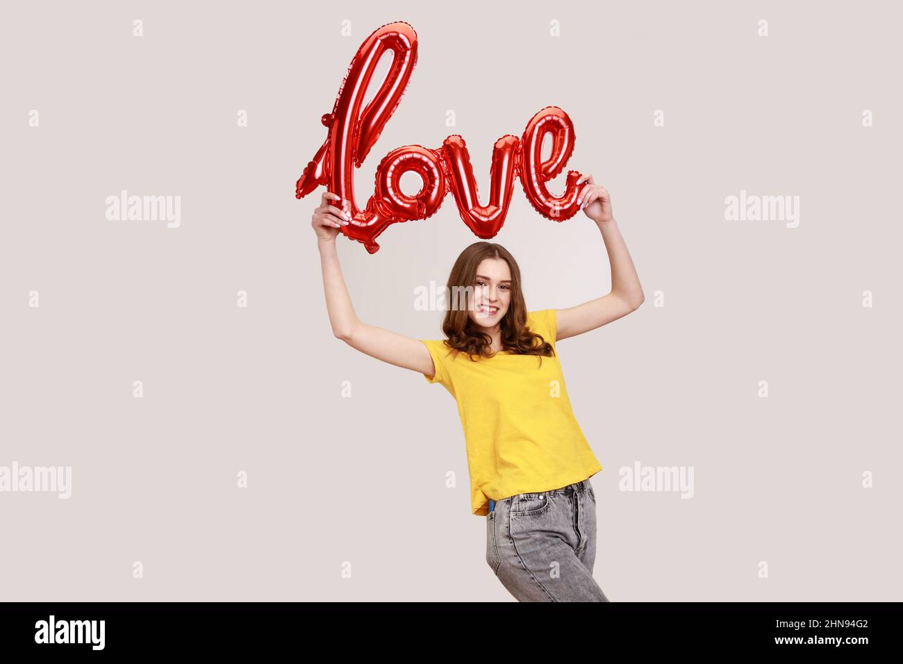 Beautiful brown haired female of young age holding balloons letters above her hear, raised arms with foil word love, wearing yellow T-shirt. Indoor studio shot isolated on gray background. Stock Photo