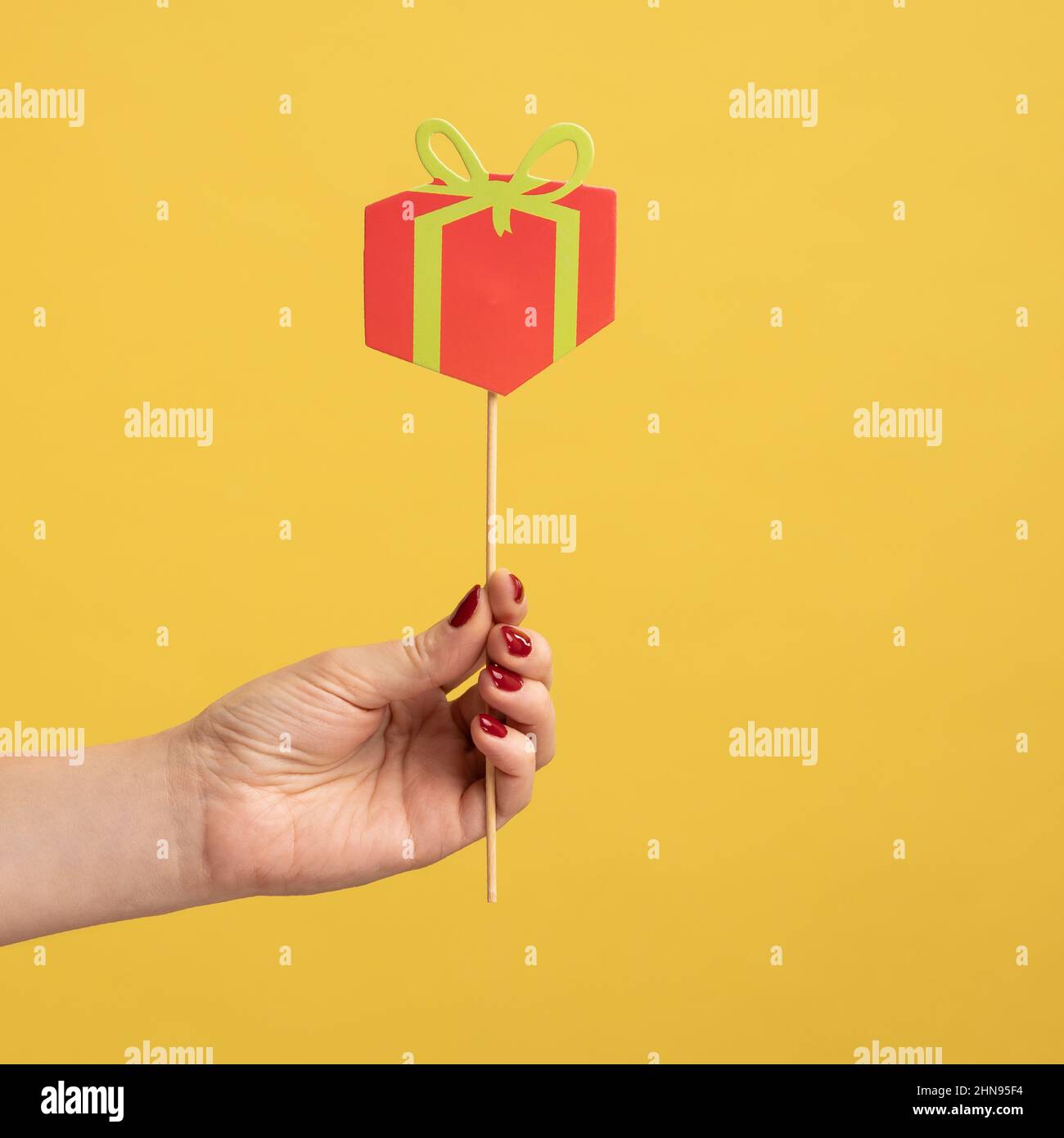 Closeup side view portrait of woman hand holding festive party props, red little paper present box on stick, celebrating holiday. Indoor studio shot isolated on yellow background. Stock Photo