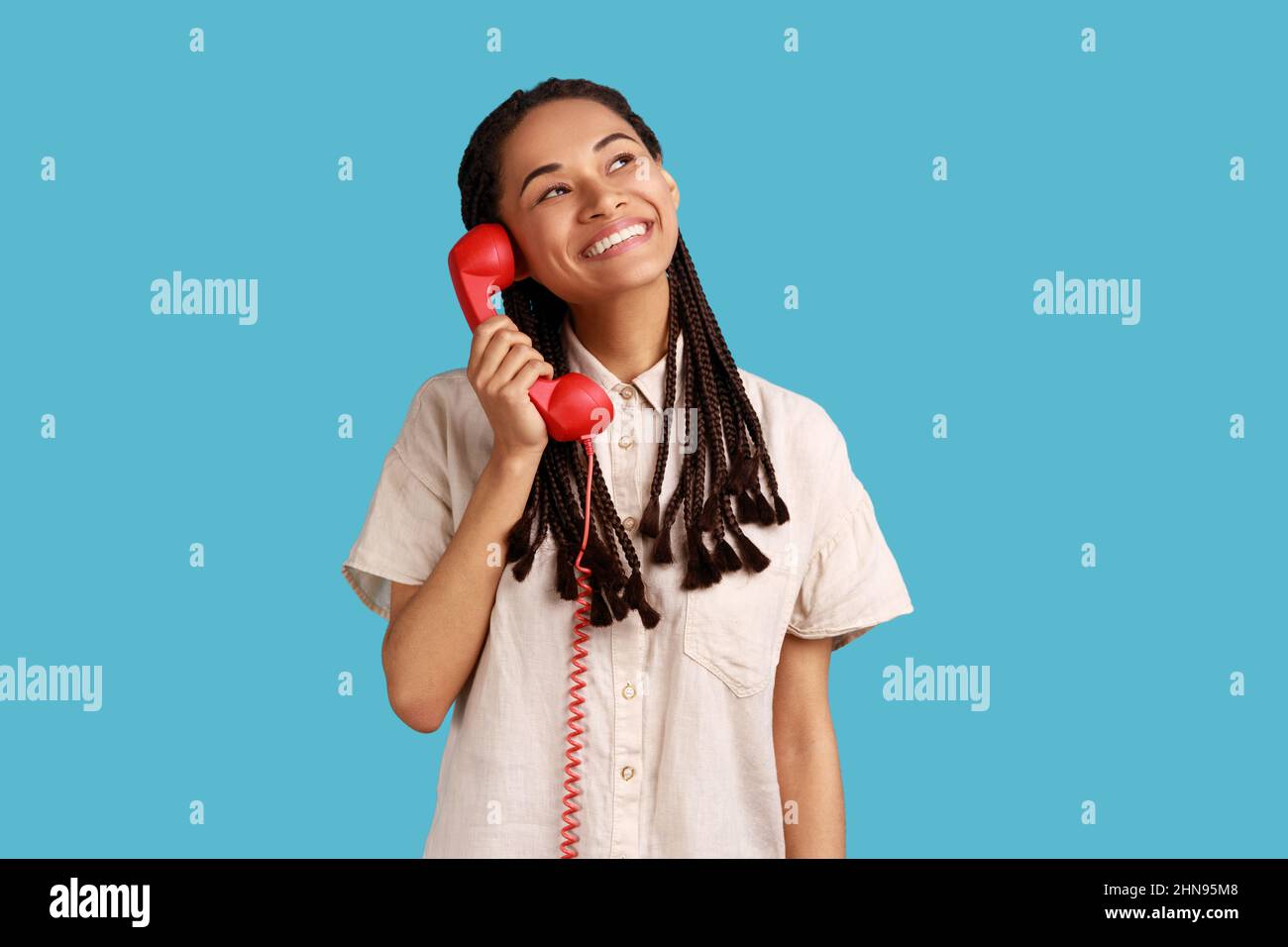 Portrait of happy smiling woman with black dreadlocks holding red retro phone in hands, looking away with dreamy facial expression, wearing white shirt. Indoor studio shot isolated on blue background. Stock Photo