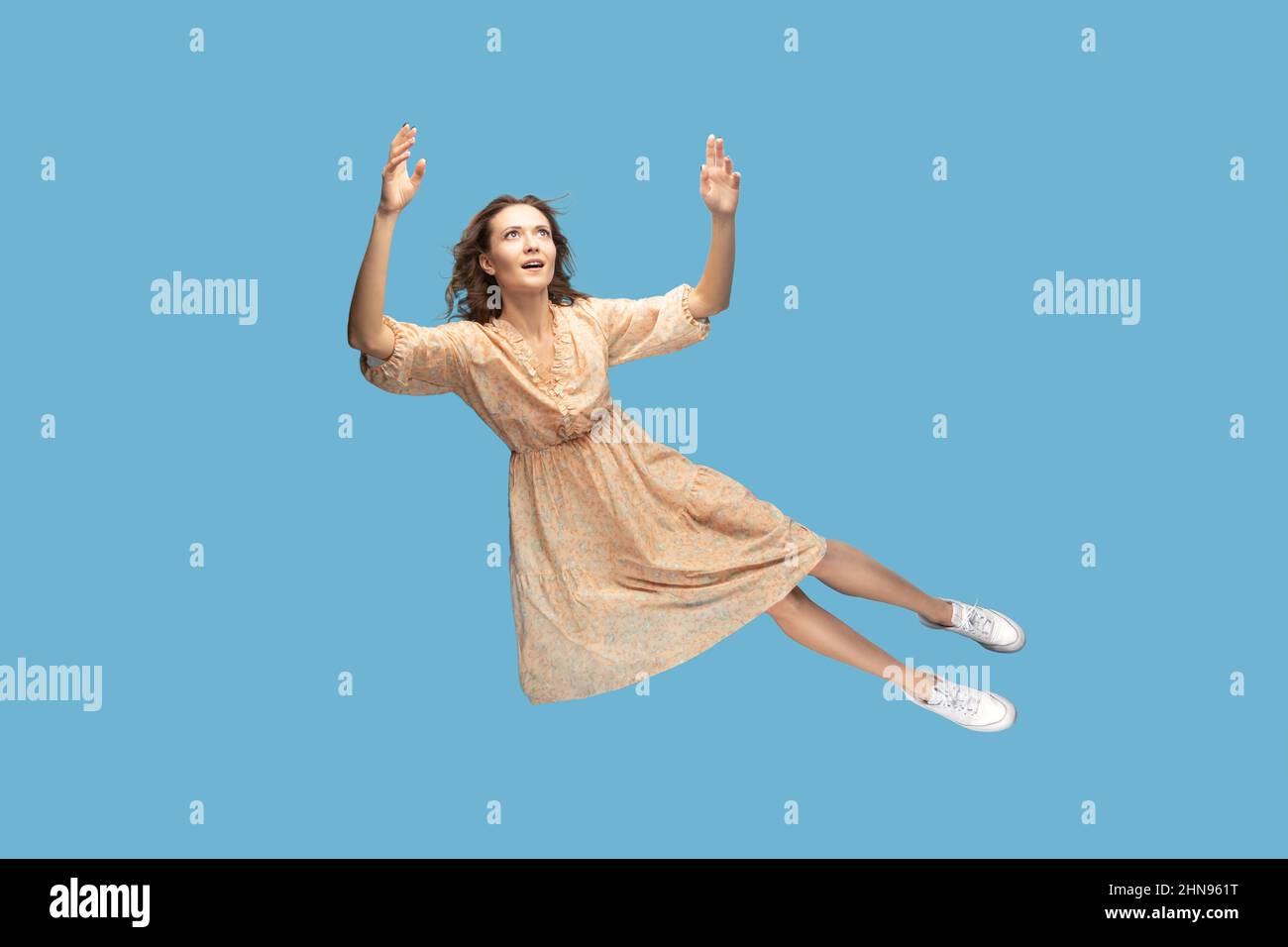 Hovering in air. Full length beautiful pensive girl in yellow dress levitating flying in mid-air, looking up with dreamy relaxed expression. indoor studio shot isolated on blue background Stock Photo