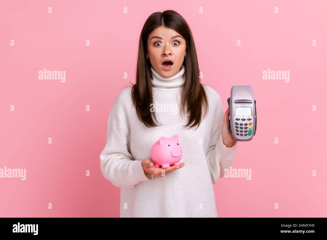 Shocked girl with dark hair showing pos payment terminal and piggybank, using cashless payments, nfc, wearing white casual style sweater. Indoor studio shot isolated on pink background. Stock Photo