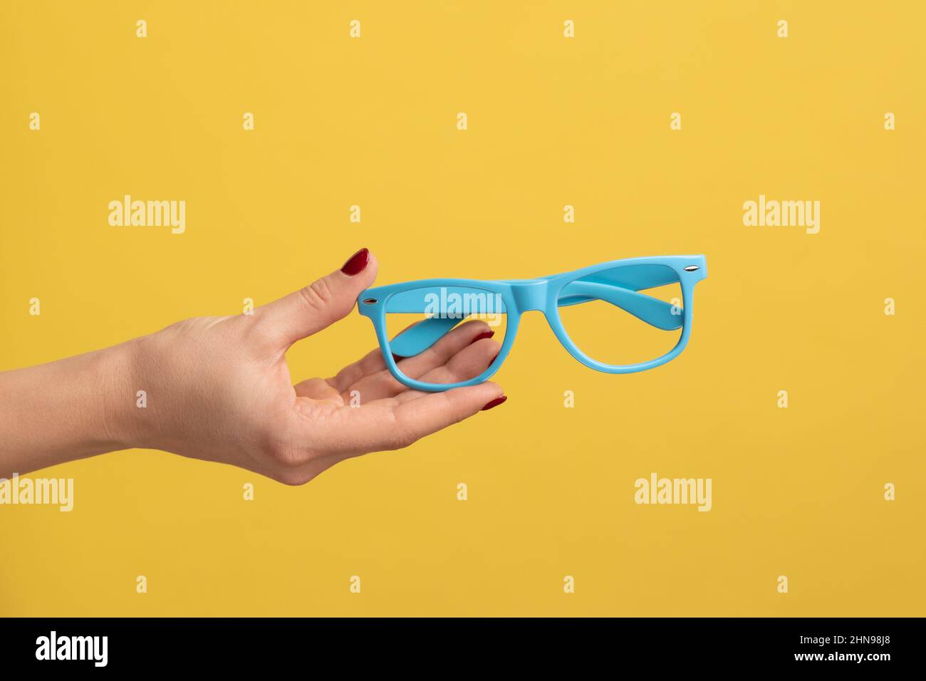 Profile side view closeup of woman hand holding and showing blue eyeglasses frame. Indoor studio shot isolated on yellow background. Stock Photo