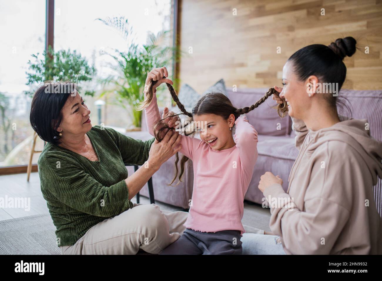 Happy little girl sitting on floor, her mother and grandmother making her plaits at home. Stock Photo