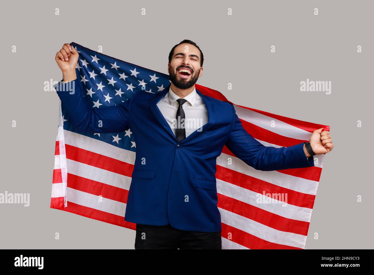 Excited bearded man holding USA flag and looking at camera with rejoice look, celebrating national holiday, wearing official style suit. Indoor studio shot isolated on gray background. Stock Photo