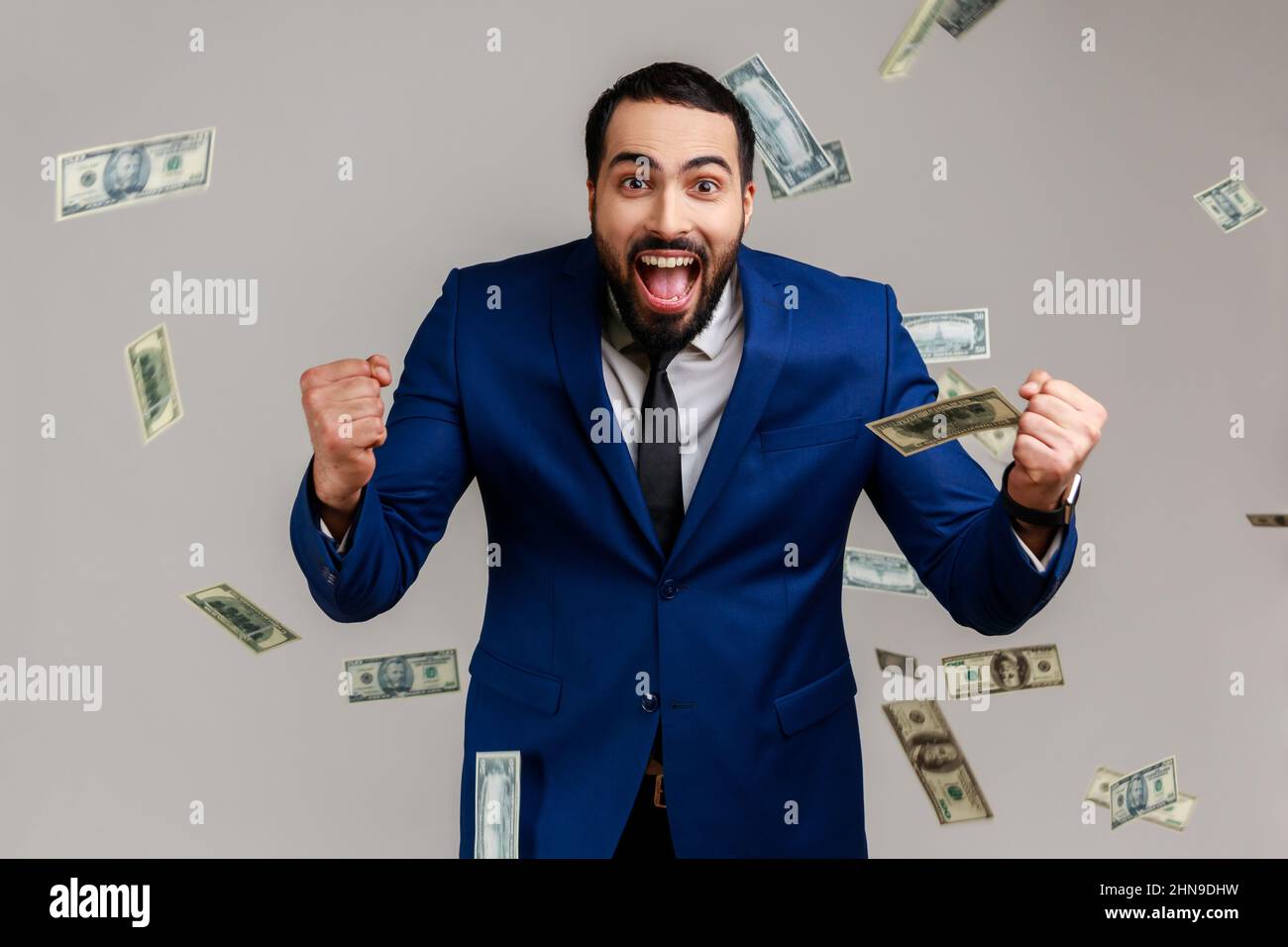 Excited bearded man standing under money with clenched fists and yelling happily, celebrating victory and richness, wearing official style suit. Indoor studio shot isolated on gray background. Stock Photo