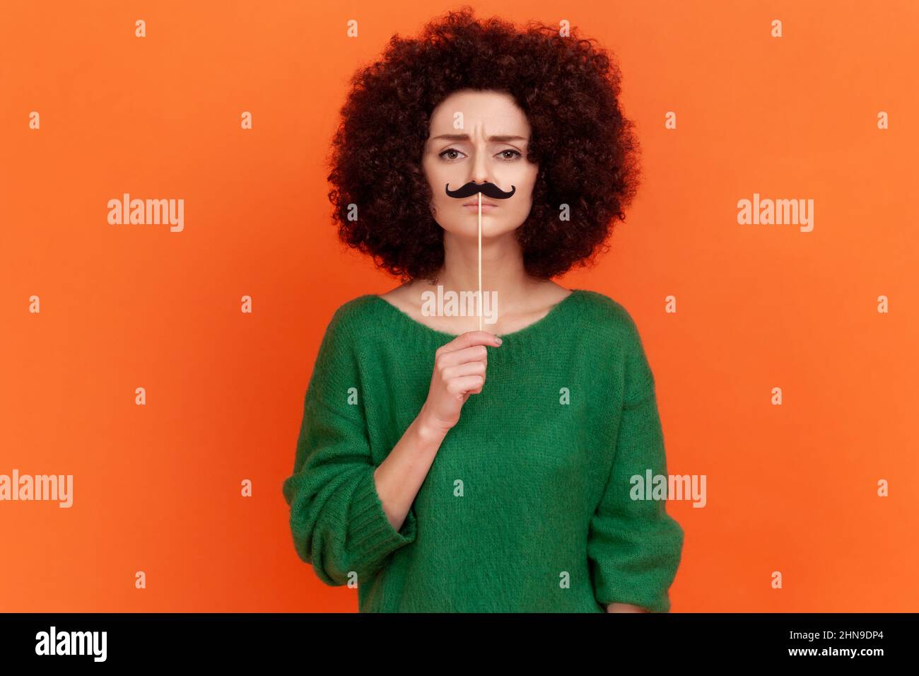 Funny woman with Afro hairstyle wearing green casual style sweater standing with paper mustache on stick, festive mood, masquerade Indoor studio shot isolated on orange background. Stock Photo