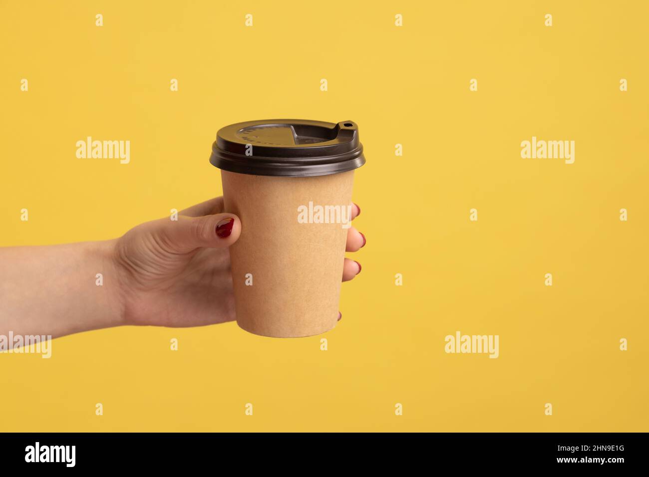 Closeup hand giving coffee in disposable cup, hot drink with caffeine to boost energy in morning, coffee break at work, tasty beverage and relaxation. Indoor studio shot isolated on yellow background. Stock Photo
