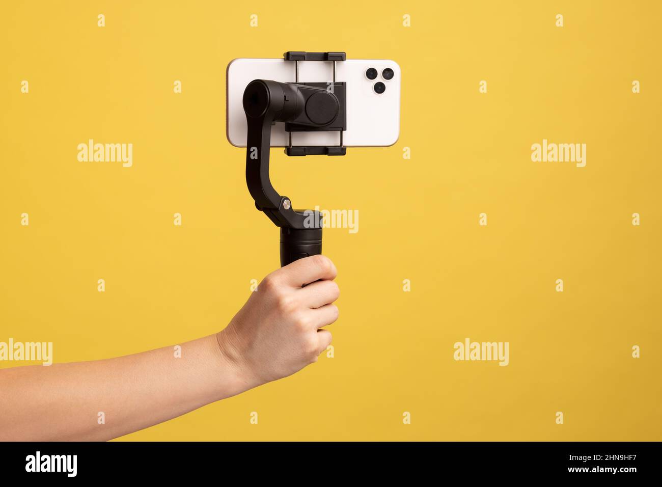 Closeup side view of woman hand holding steadicam with phone, for making video or has livestream. Indoor studio shot isolated on yellow background. Stock Photo
