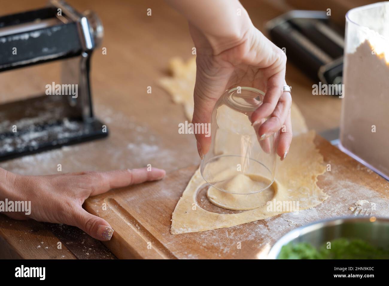 Woman's hand stands out with a small water glass for dumplings from dough rounds on a wooden board in the kitchen Stock Photo