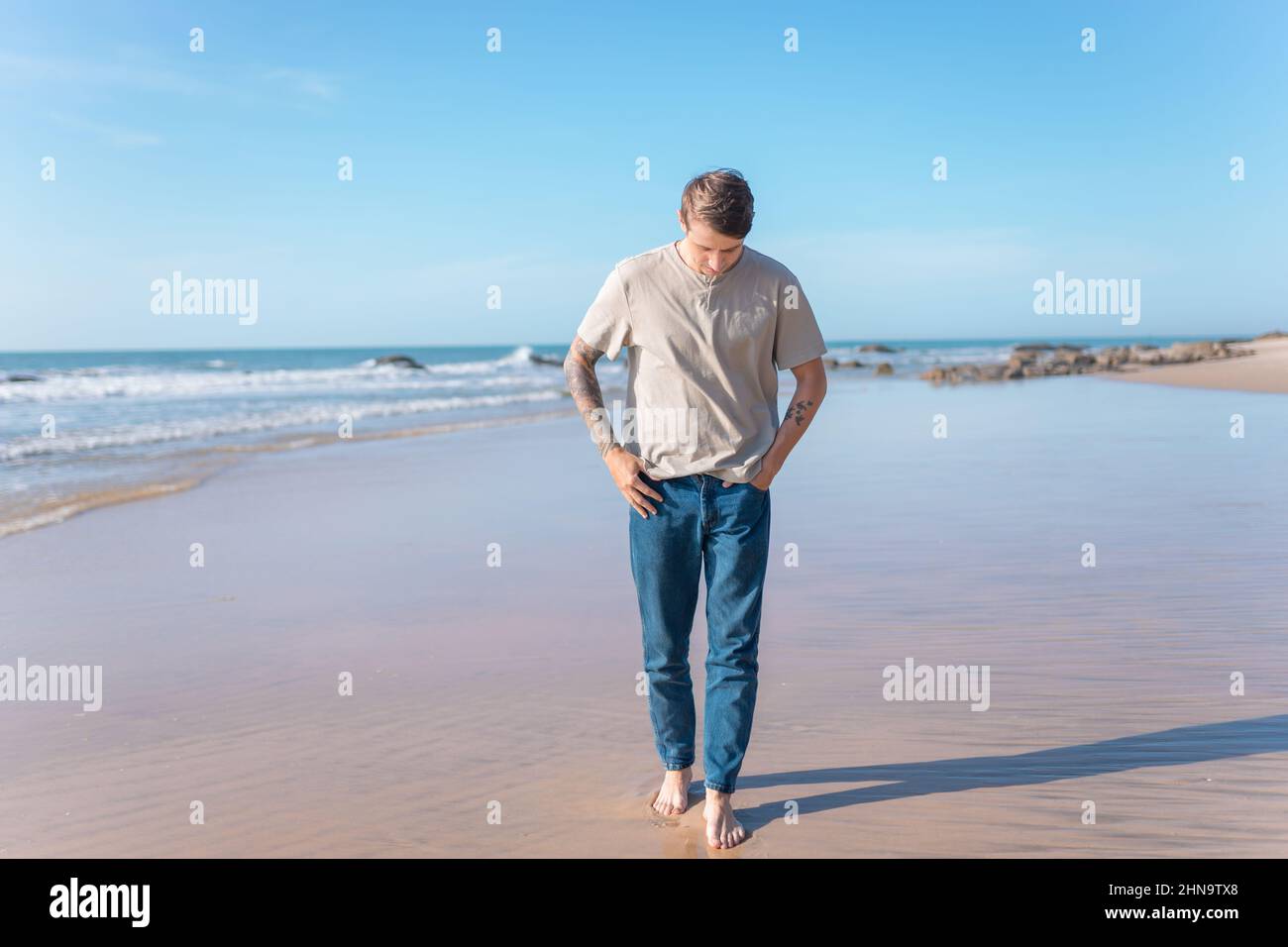 Attractive young caucasian man with full-arm tattoo in casual clothing walking on the beach barefoot. Scenic ocean view, morning. High quality photo Stock Photo