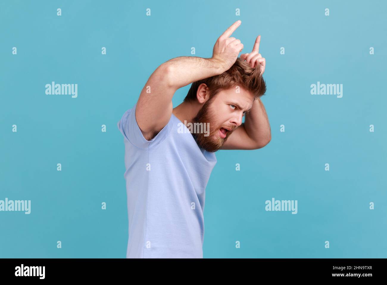 Side view of angry bully bearded man showing bull horn gesture with fingers over head, looking hostile and threatening, aggressive face. Indoor studio shot isolated on blue background. Stock Photo
