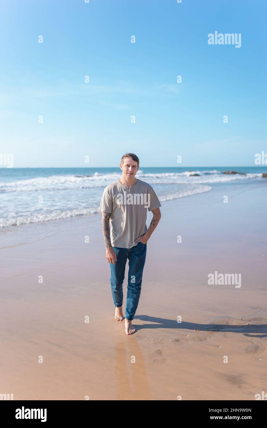 Attractive young caucasian man with full-arm tattoo in casual clothing walking on the beach barefoot. Scenic ocean view, morning. High quality photo Stock Photo
