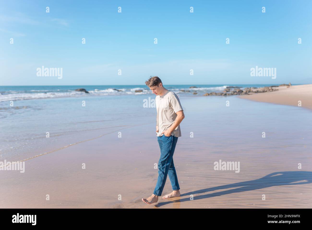Attractive young caucasian man with full-arm tattoo in casual clothing walking on the beach barefoot. Scenic ocean view, morning. High quality photo Stock Photo