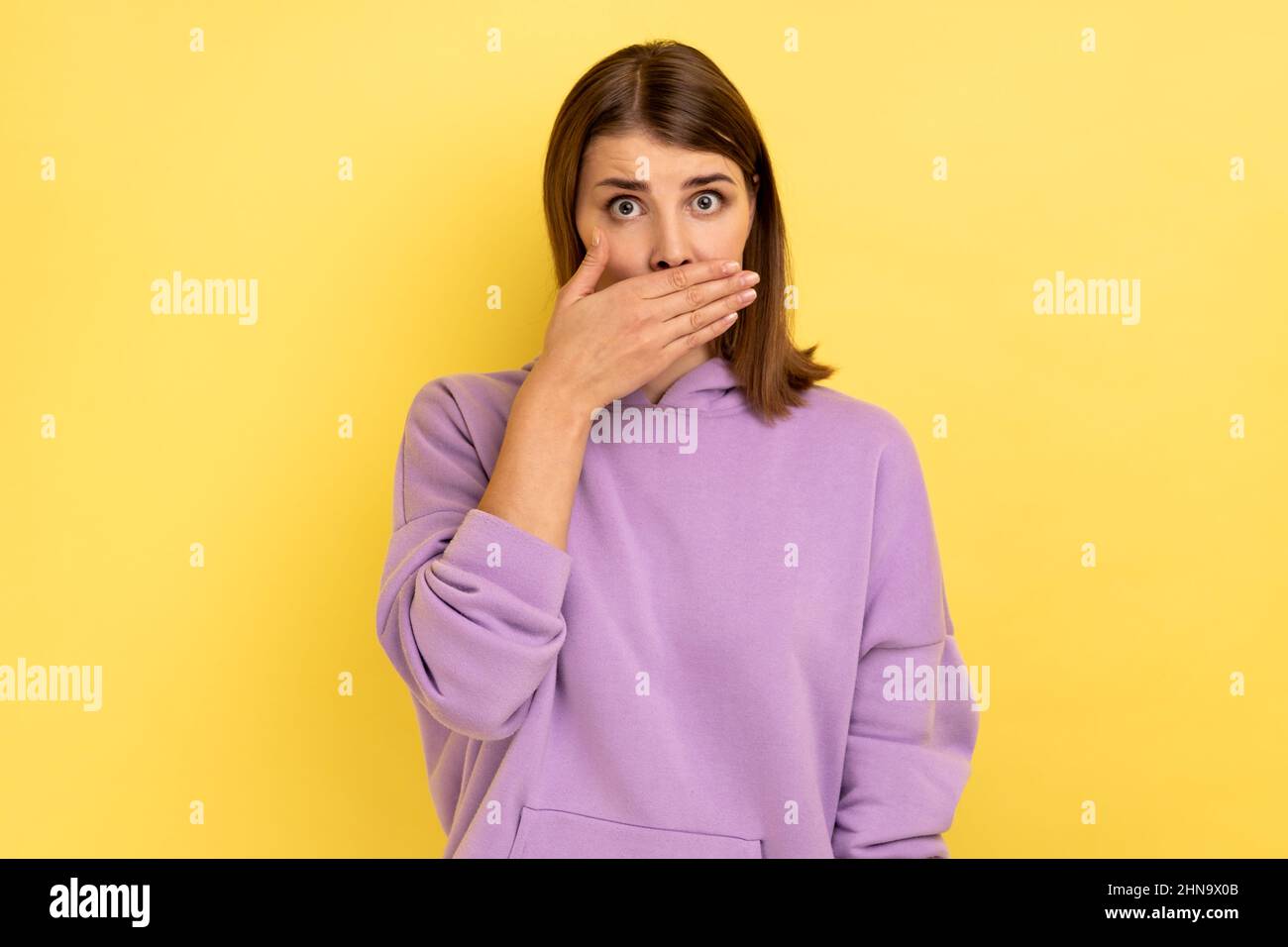 Portrait of shocked dark haired woman put hand on mouth and looking with fear in her eyes, keeping terrible secret, wearing purple hoodie. Indoor studio shot isolated on yellow background. Stock Photo