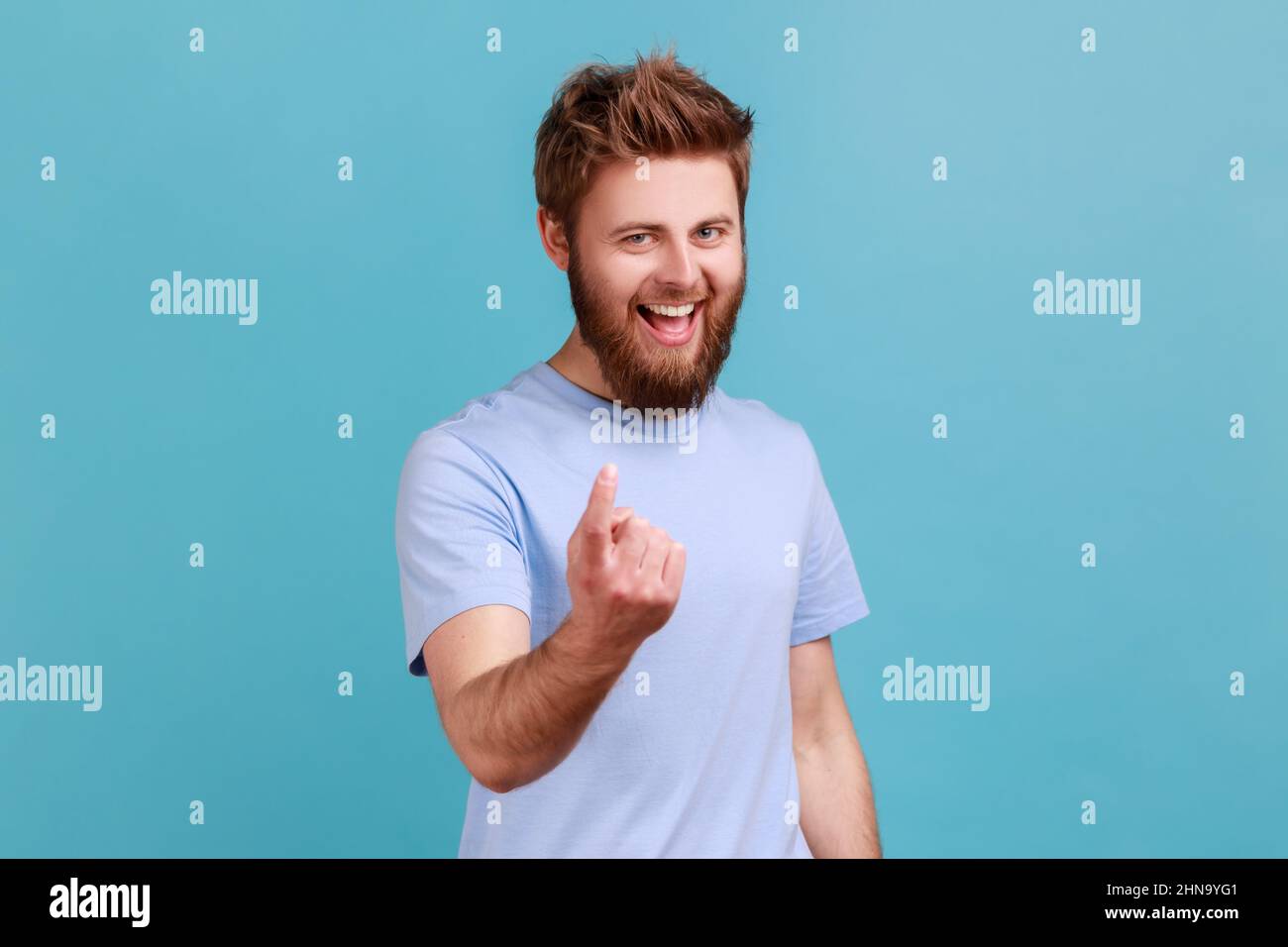 Come here. Portrait of bearded man making beckoning gesture, inviting to come, flirting and looking playful, having excited facial expression. Indoor studio shot isolated on blue background. Stock Photo