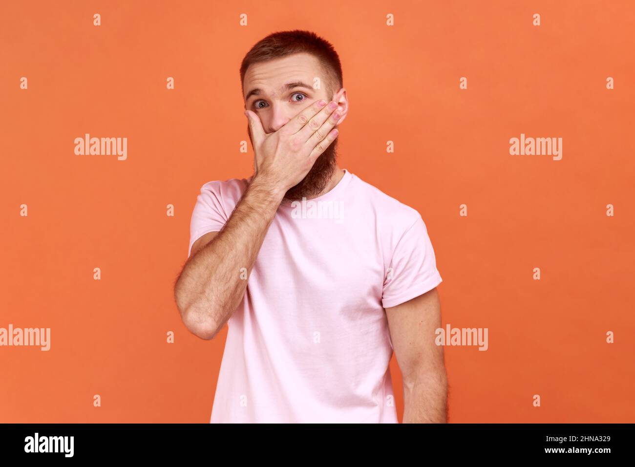 Portrait of bearded man covering mouth with hand to keep silent, afraid to say secret, looking with intimidated expression, wearing pink T-shirt. Indoor studio shot isolated on orange background. Stock Photo