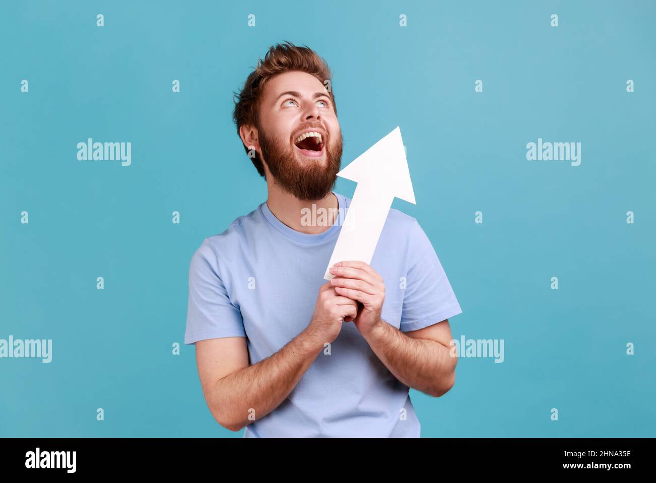 Portrait of excited positive optimistic bearded man holding arrow pointing up looking at camera with smile, growth and increase concept. Indoor studio shot isolated on blue background. Stock Photo