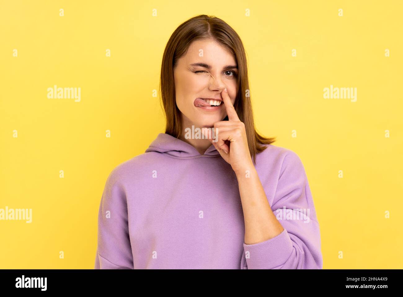 Crazy weird woman with brown hair holding finger in her nose showing tongue, uncultured bored girl having fun, bad manners, wearing purple hoodie. Indoor studio shot isolated on yellow background. Stock Photo