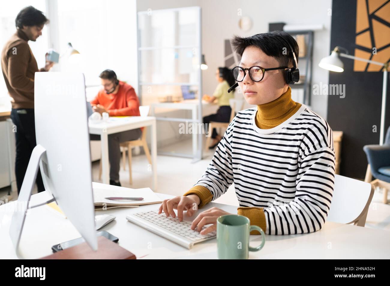 Asian operator in headphones sitting at the table with computer and typing on keyboard, she working with clients online Stock Photo