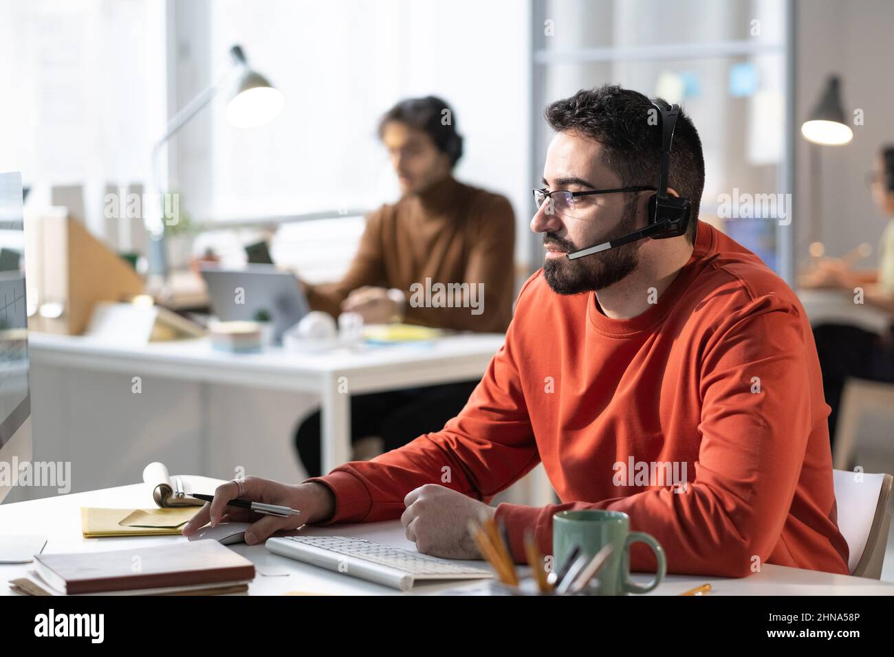 Young bearded man in headphones speaking to the microphone and talking to the clients while using computer at the table at office Stock Photo