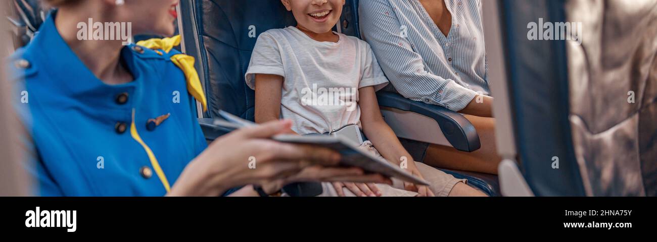 Female air hostess trying to entertain a kid on the plane by offering a book to read. Cabin crew provide service to family in airplane Stock Photo
