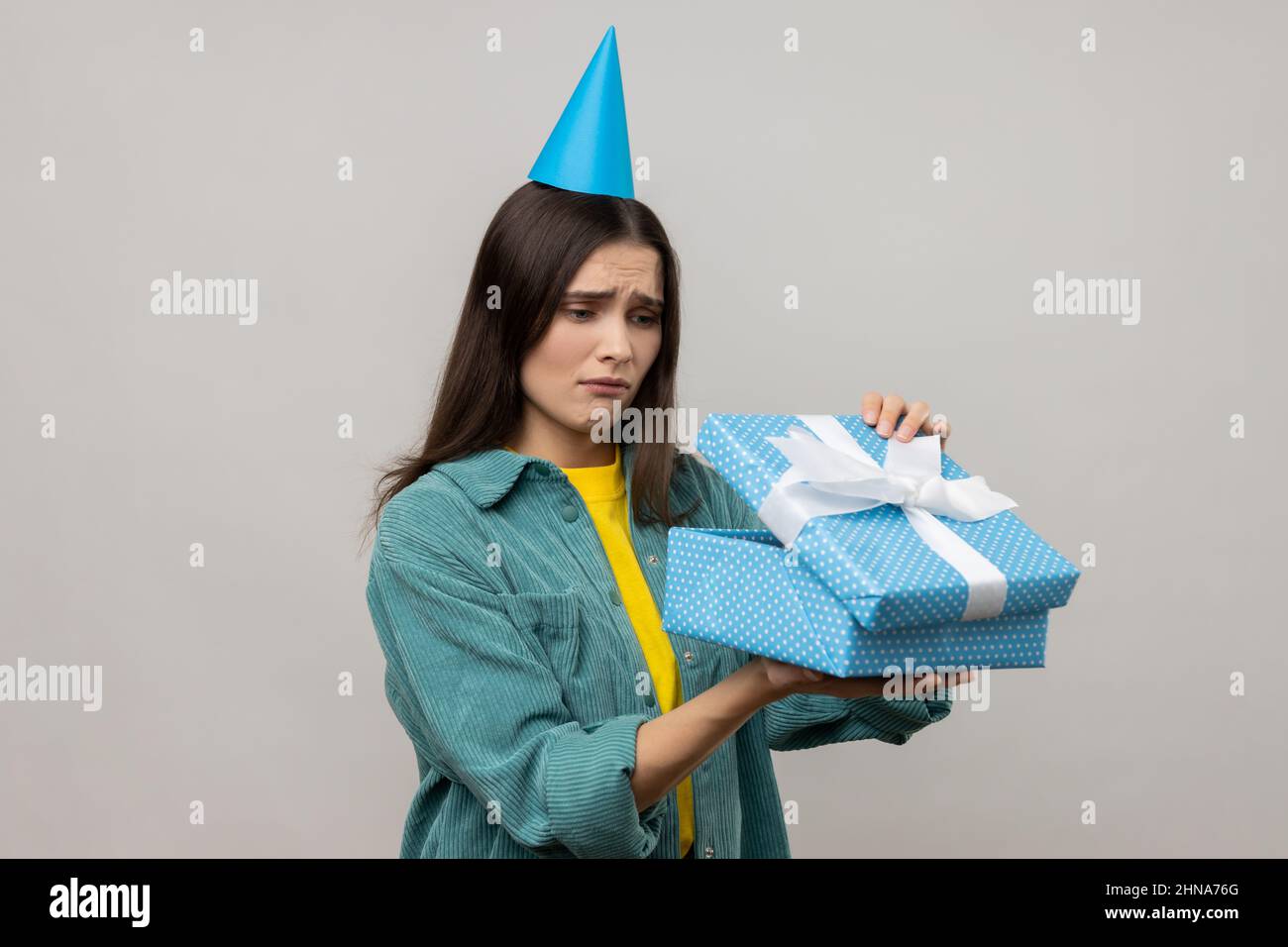 Portrait of upset woman opening birthday gift box and looking inside with disappointed expression, unwrapping bad present, wearing casual style jacket. Indoor studio shot isolated on gray background. Stock Photo