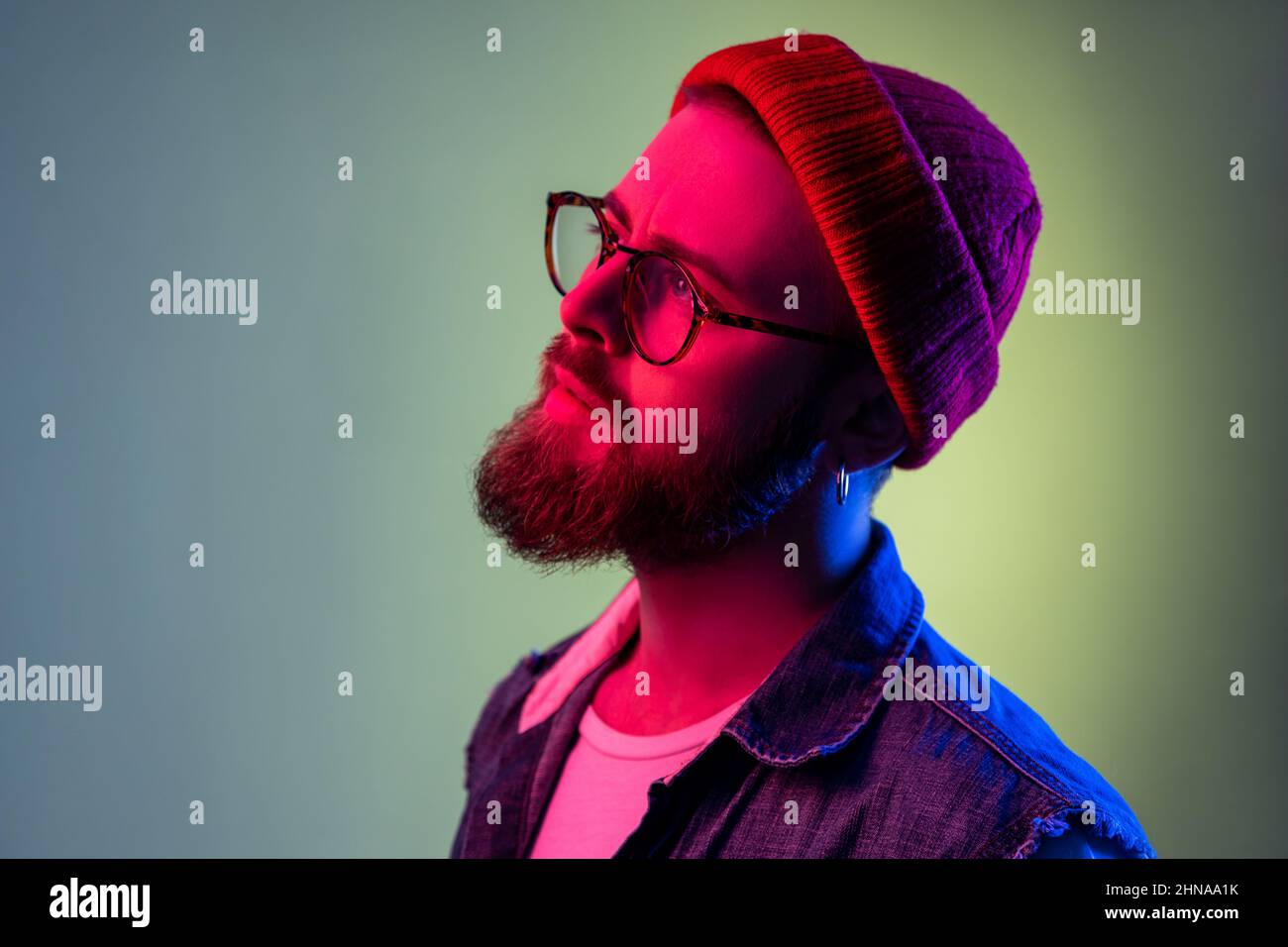 Portrait of handsome bearded hipster man in glasses, looking away with dreamy expression, wearing red beanie hat, beauty and fashion. Indoor studio shot isolated on colorful neon light background. Stock Photo
