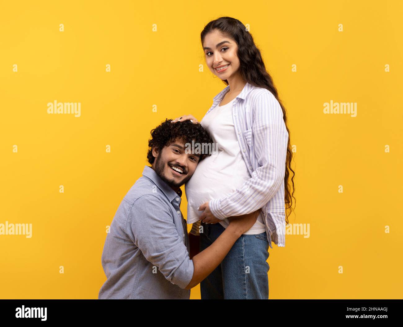 Caring indian man putting his ear on wife's pregnant tummy, listening to baby's heartbeat, yellow studio background Stock Photo