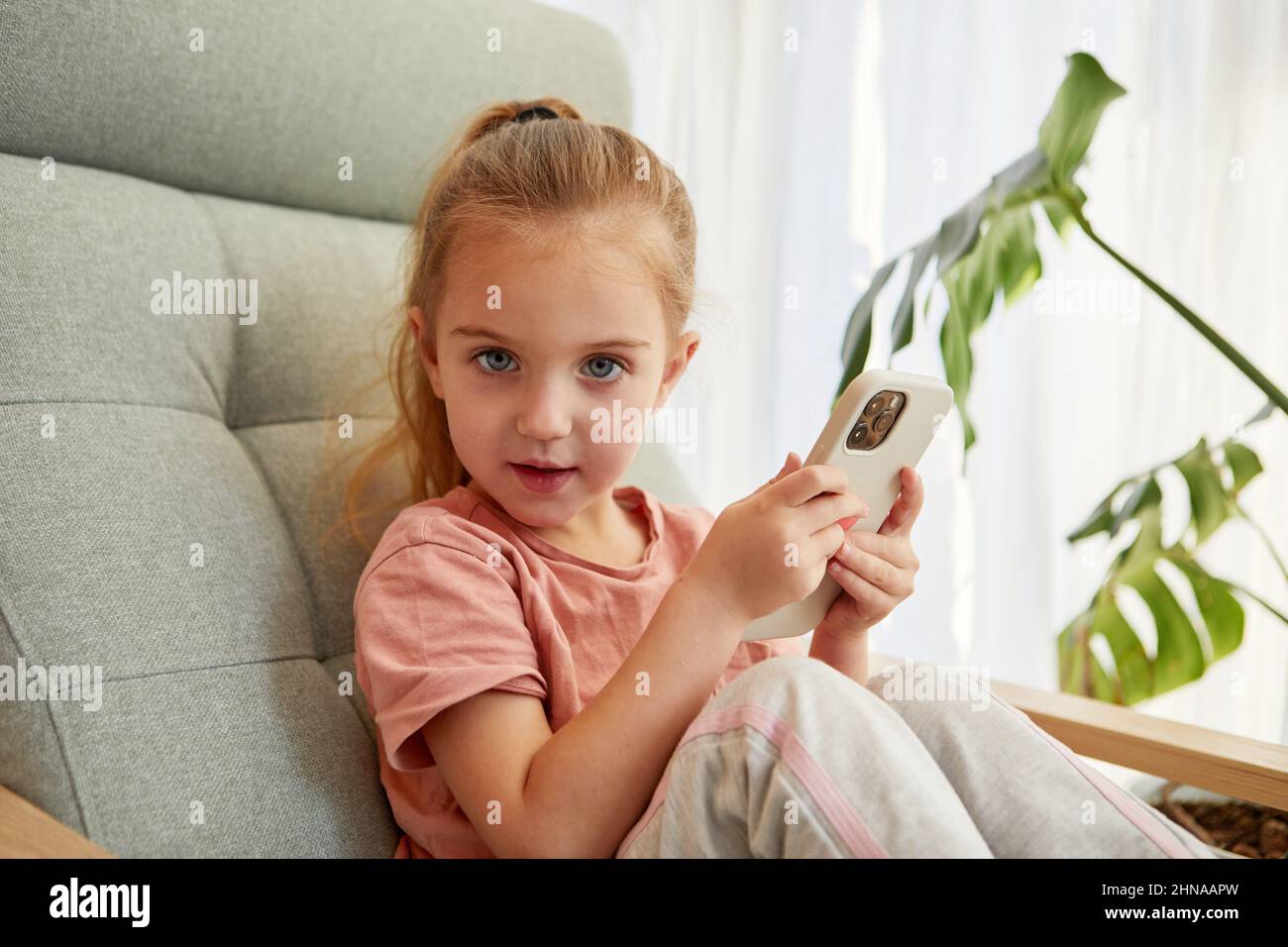 Portrait of reproachful little girl holding mobile phone and looking at camera sitting on chair at home Stock Photo